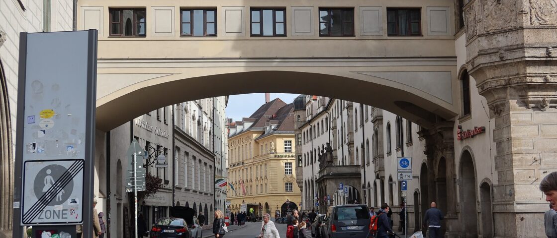 An adult pushes a covered stroller across Munich's Sparkassenstrasse beneath the enclosed bridge between historic buildings.