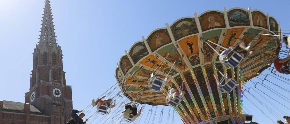 A chair swing ride in full spin beside the red-brick spire of the Mariahilfkirche under a blue sky