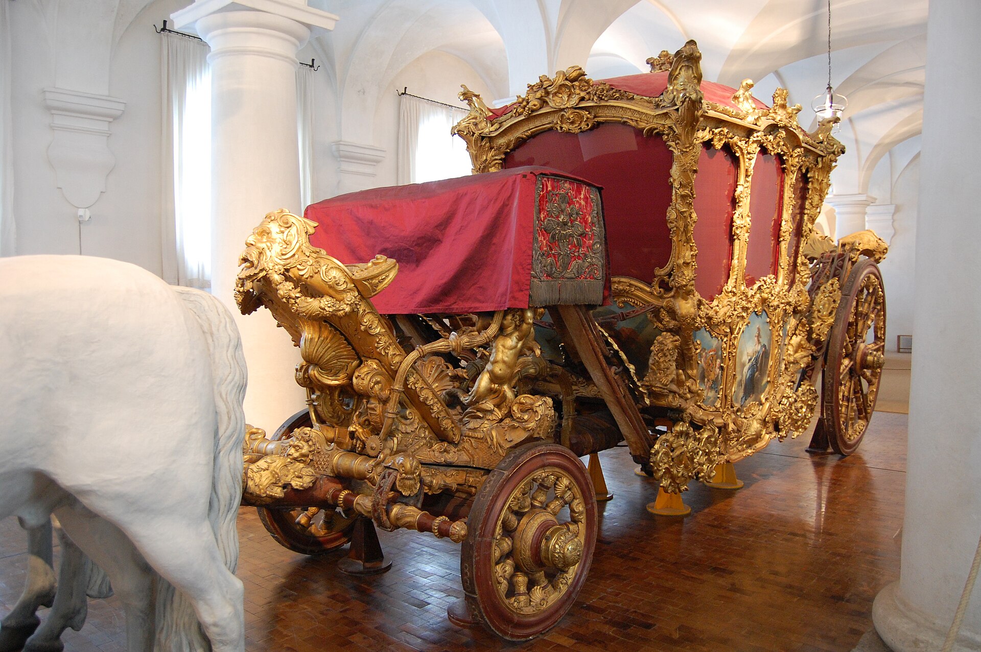 Nymphenburg Palace Munich: Complete Visitor Guide (2026) 4 A gilded royal coach with red upholstery and sculpted figures stands beside a model horse in Munich’s Marstallmuseum.