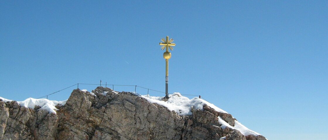 The golden summit cross stands above snow patches on the rocky crest of the Zugspitze beneath a clear blue sky.