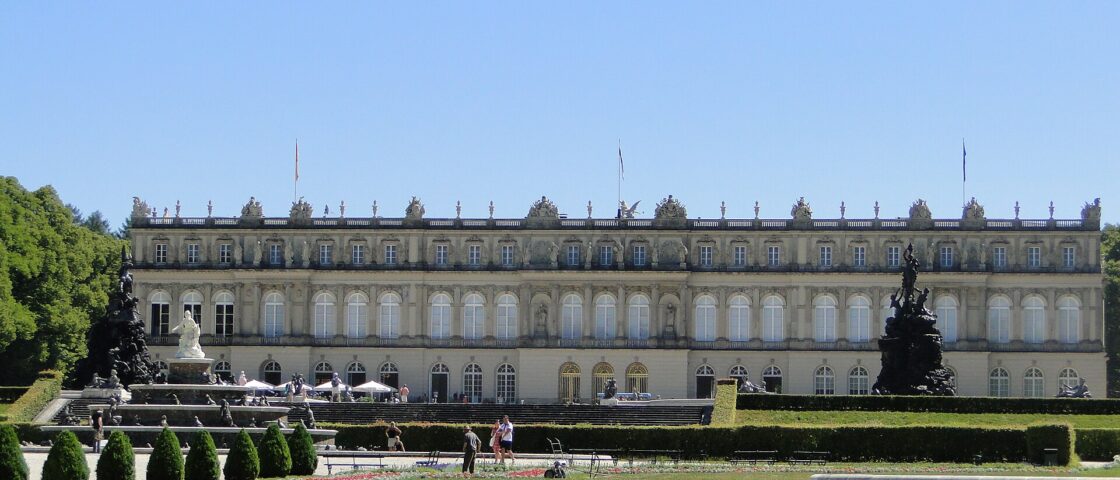 Herrenchiemsee New Palace stretches behind sculpted fountains, trimmed hedges and flower beds, with visitors walking through the formal garden.