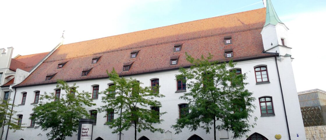 Munich City Museum’s white facade and steep red roof face the street, with trees and bicycles along the pavement.
