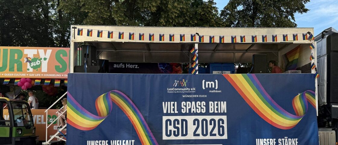 A parade truck with a sound stage and a banner reading Viel Spass beim CSD 2026 with rainbow stripes, under trees
