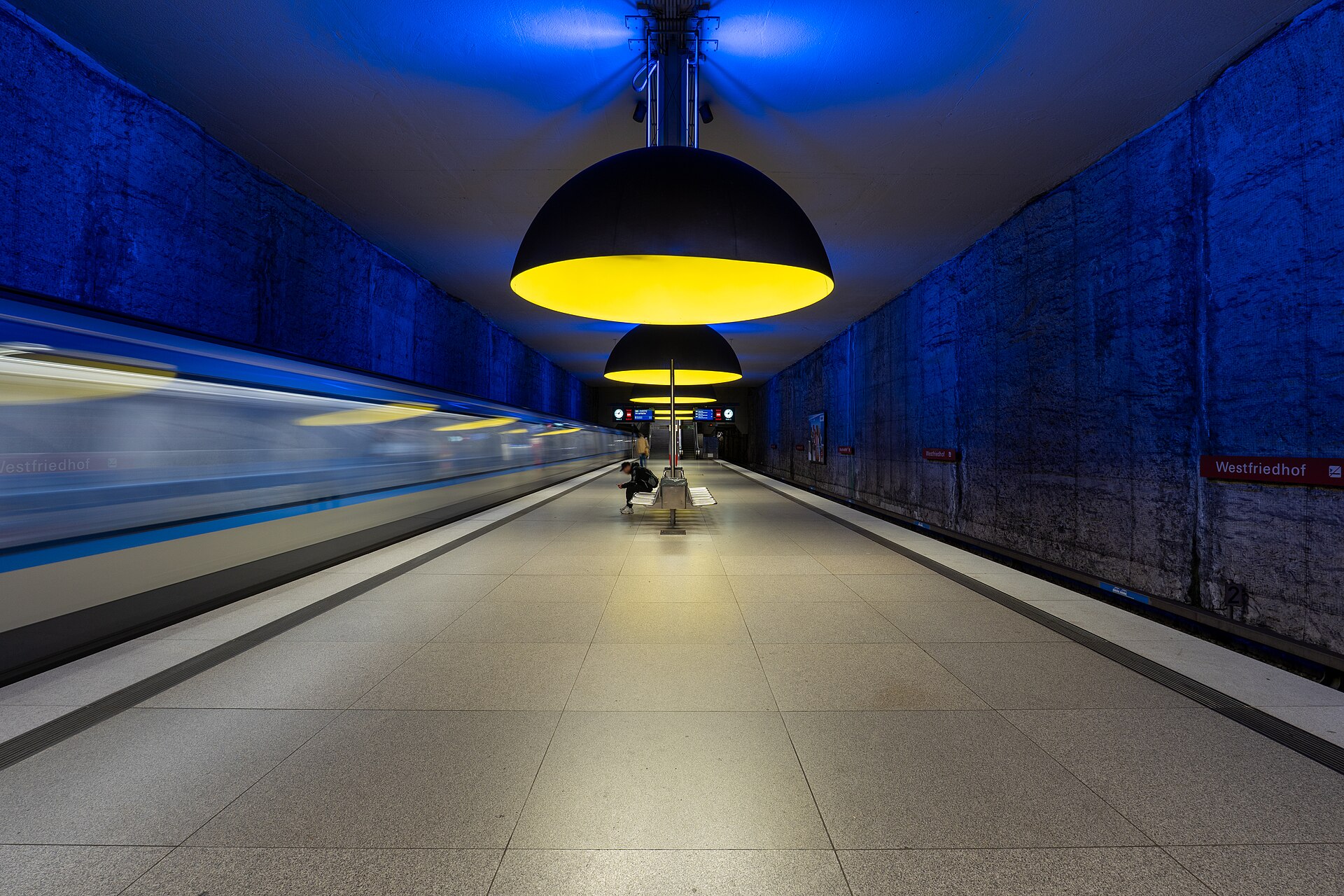 Getting Around Munich: Complete Transport Guide 1 A Munich U-Bahn train passes Westfriedhof station beneath oversized yellow lamps, beside blue lit walls and an island platform.
