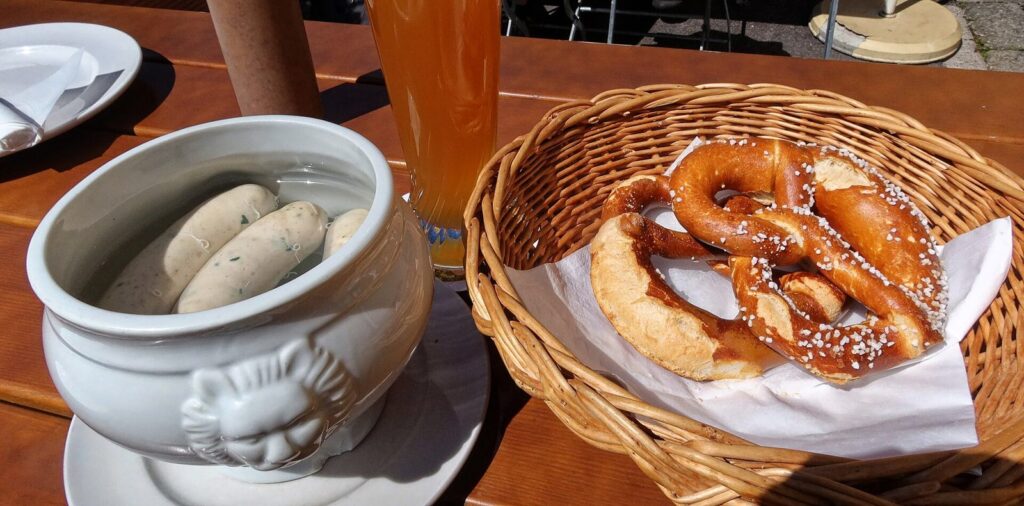 Weisswurst in a white porcelain tureen beside a basket of pretzels and a wheat beer