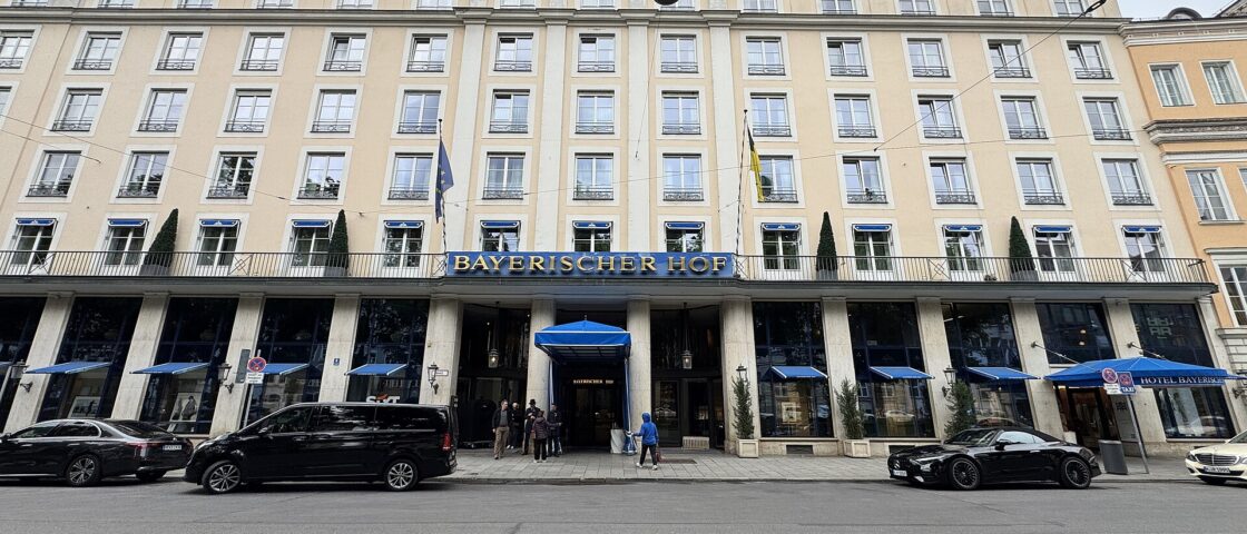 Munich’s Bayerischer Hof faces Promenadeplatz with a pale facade, blue entrance canopy, flags and passing cars.