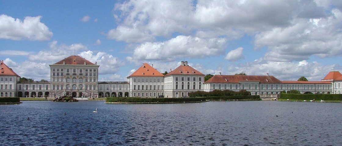 Nymphenburg Palace in Munich stretches beside a broad basin, with red-roofed pavilions and waterbirds beneath a cloud-filled sky.