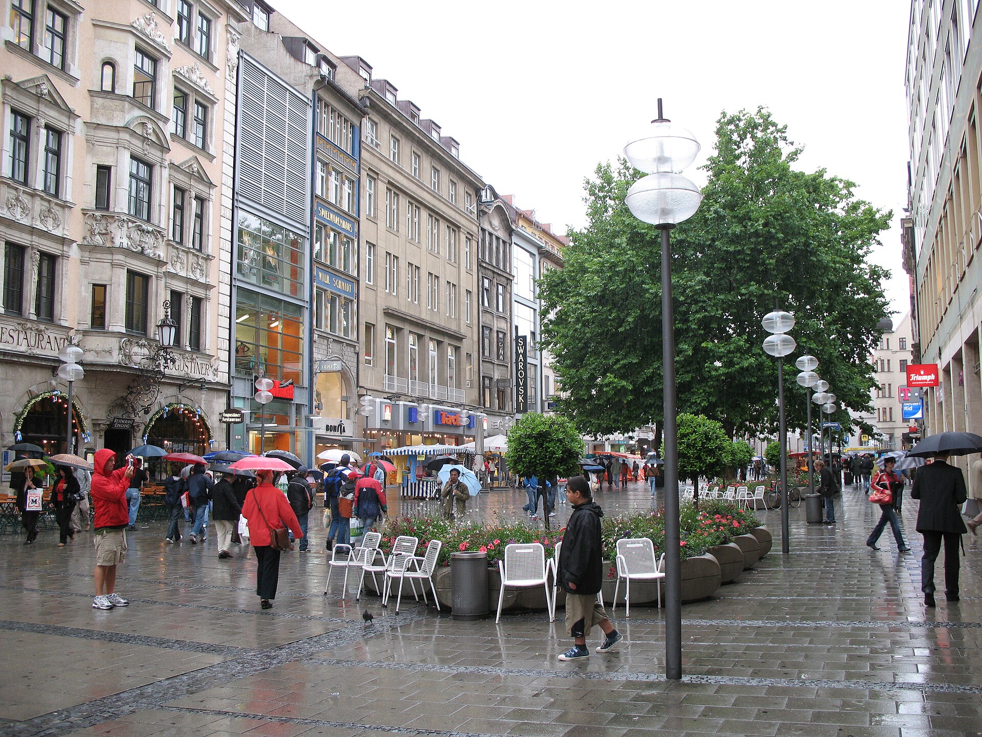 Getting Around Munich: Complete Transport Guide 7 People carrying umbrellas walk along Munich's Neuhauser Strasse past outdoor chairs, planters and shops on rain-darkened paving.