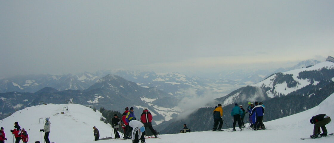 Skiers prepare their equipment on a snowy piste at Sudelfeld, with forested ridges and snow-covered Alpine peaks in the distance.