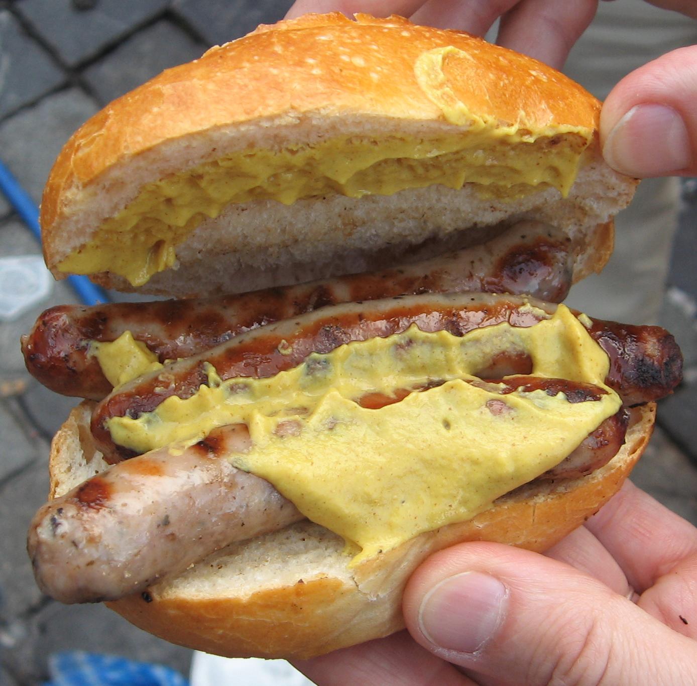 Nuremberg Day Trip from Munich: Complete History and Food Guide 2 Three small grilled Nuremberg sausages with yellow mustard fill a crusty bread roll held open in two hands.
