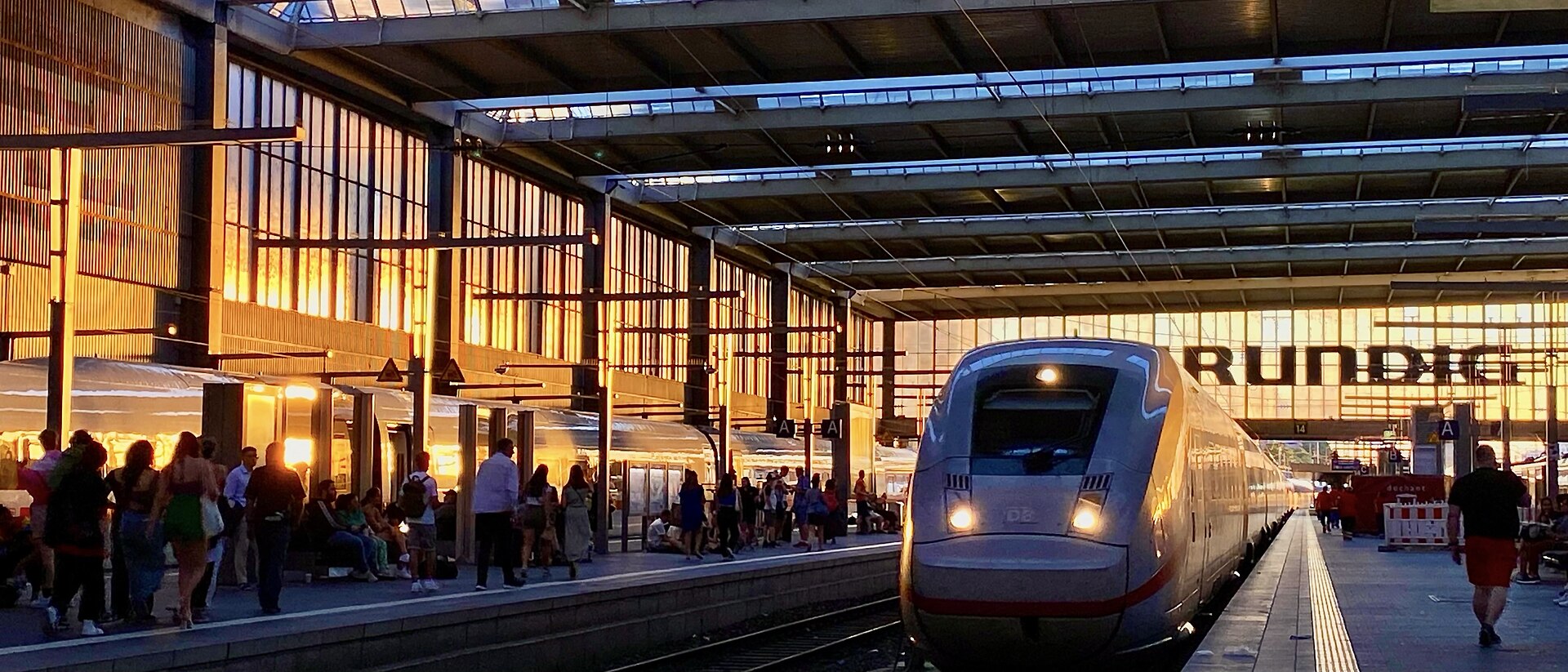 Getting Around Munich: Complete Transport Guide 2 A train arrives beside waiting passengers under the sunlit steel and glass roof of Munich Hauptbahnhof.