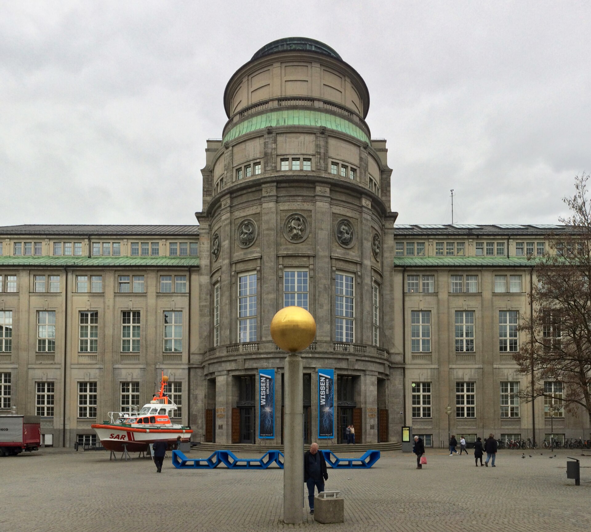 Munich Museum Pass and Discount Tickets: Complete 2026 Guide 4 Visitors cross the Deutsches Museum courtyard in Munich beneath a rounded tower, beside a golden sphere and rescue boat.